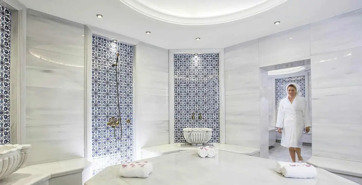 White marble hammam with blue patterned tile panels, hand shower, carved basin and towels on the navel stone
