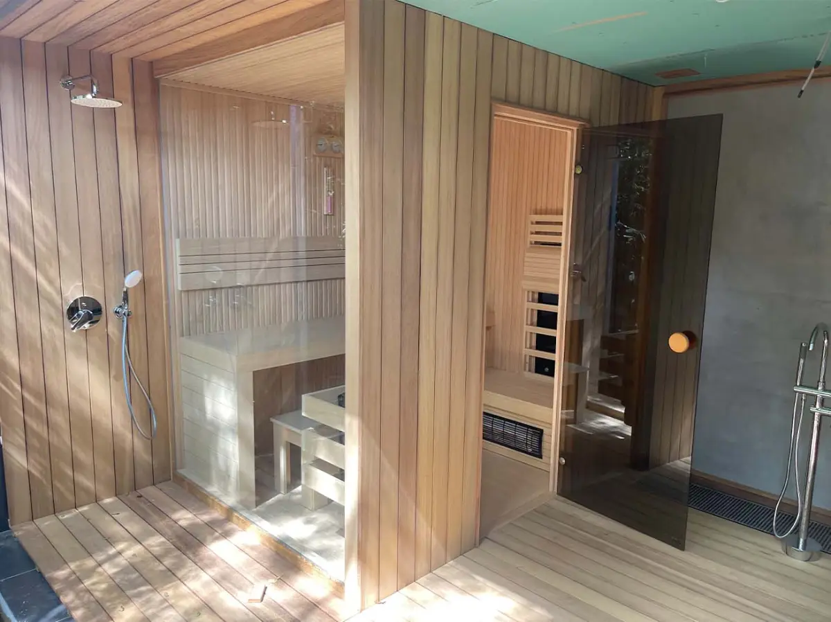 Two glass-fronted timber sauna cabins side by side on a wooden deck with rain shower