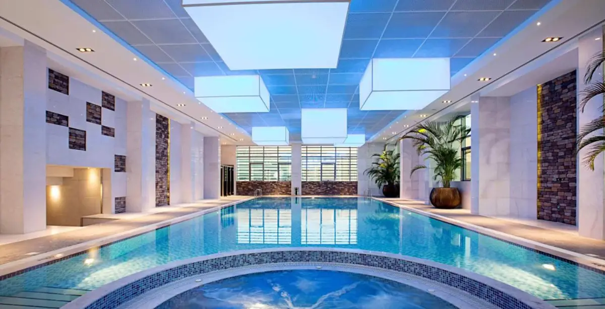 Long indoor pool with backlit blue ceiling panels, stone-clad piers and a round mosaic spa pool