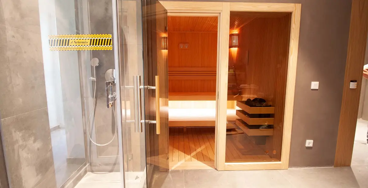Timber sauna cabin with glass door beside a walk-in shower in a grey-tiled room