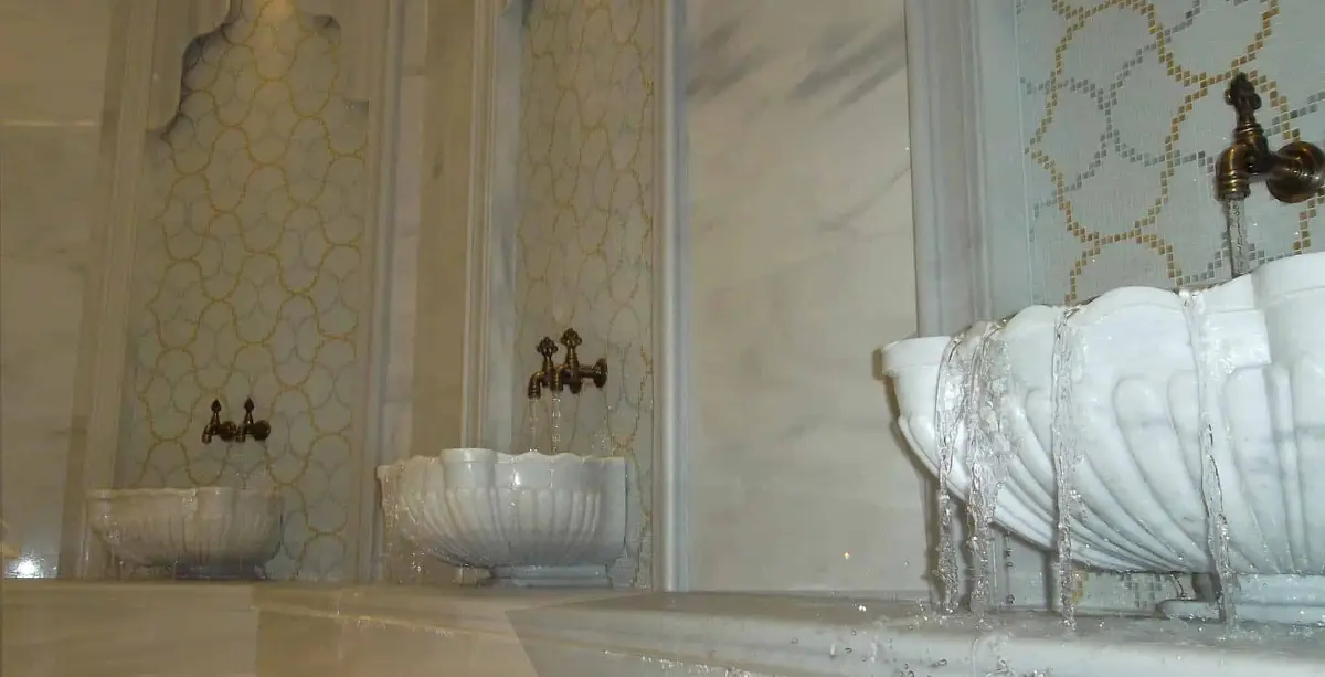 Carved white marble basins overflowing with water beneath brass taps set in gold mosaic panels