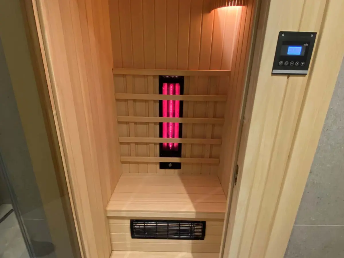 Infrared cabin with slatted backrest, glowing red emitter, floor vent and wall-mounted digital controller