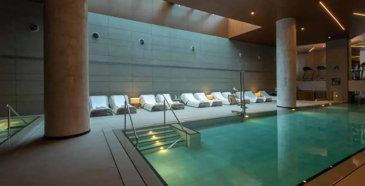 Indoor pool with lit steps, concrete columns, wall jets and a row of white loungers