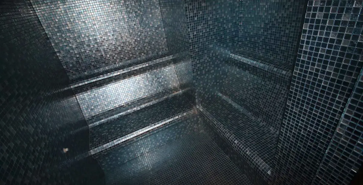 Steam room lined throughout in dark blue-grey glass mosaic, with tiered stepped benching on one side, an L-shaped bench along the corner and light strips recessed under the bench noses